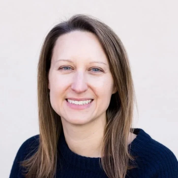 A woman with straight, shoulder-length brown hair and blue eyes, smiling in front of a plain white background, wearing a dark blue sweater.