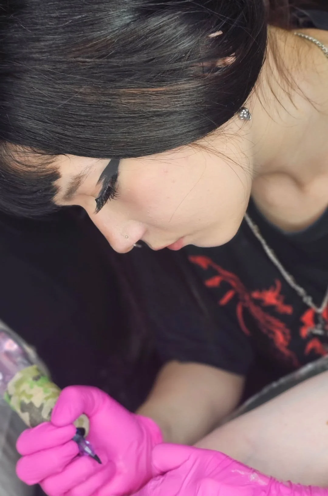 Girl with long black hair tattoing a customer with pink gloves @catarina.ink
