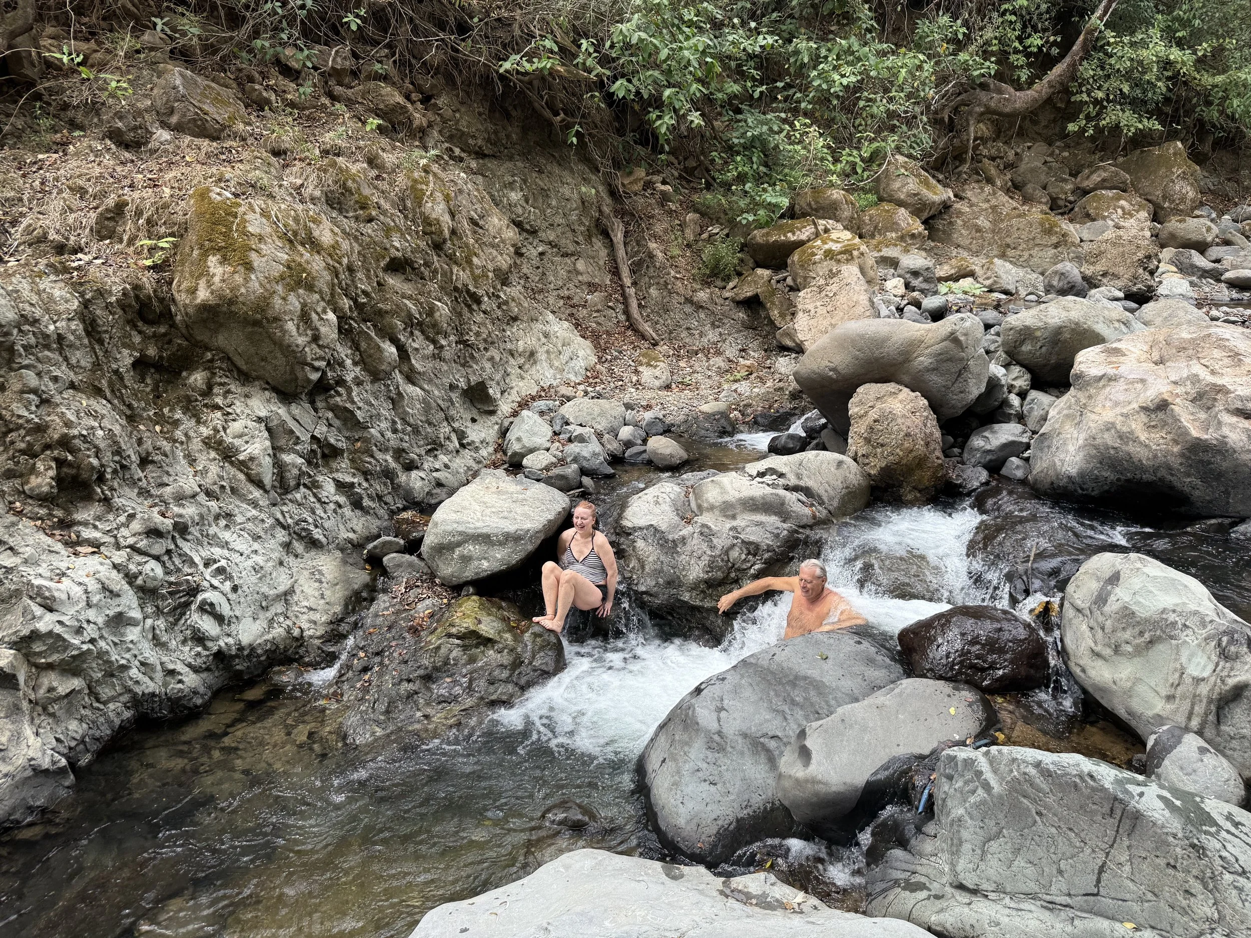 Two people, one woman and one man, sitting on rocks in a river, surrounded by large boulders and trees.