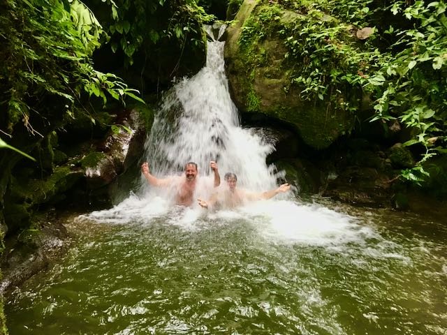 Two people swimming and enjoying a small waterfall in a lush, green forest.