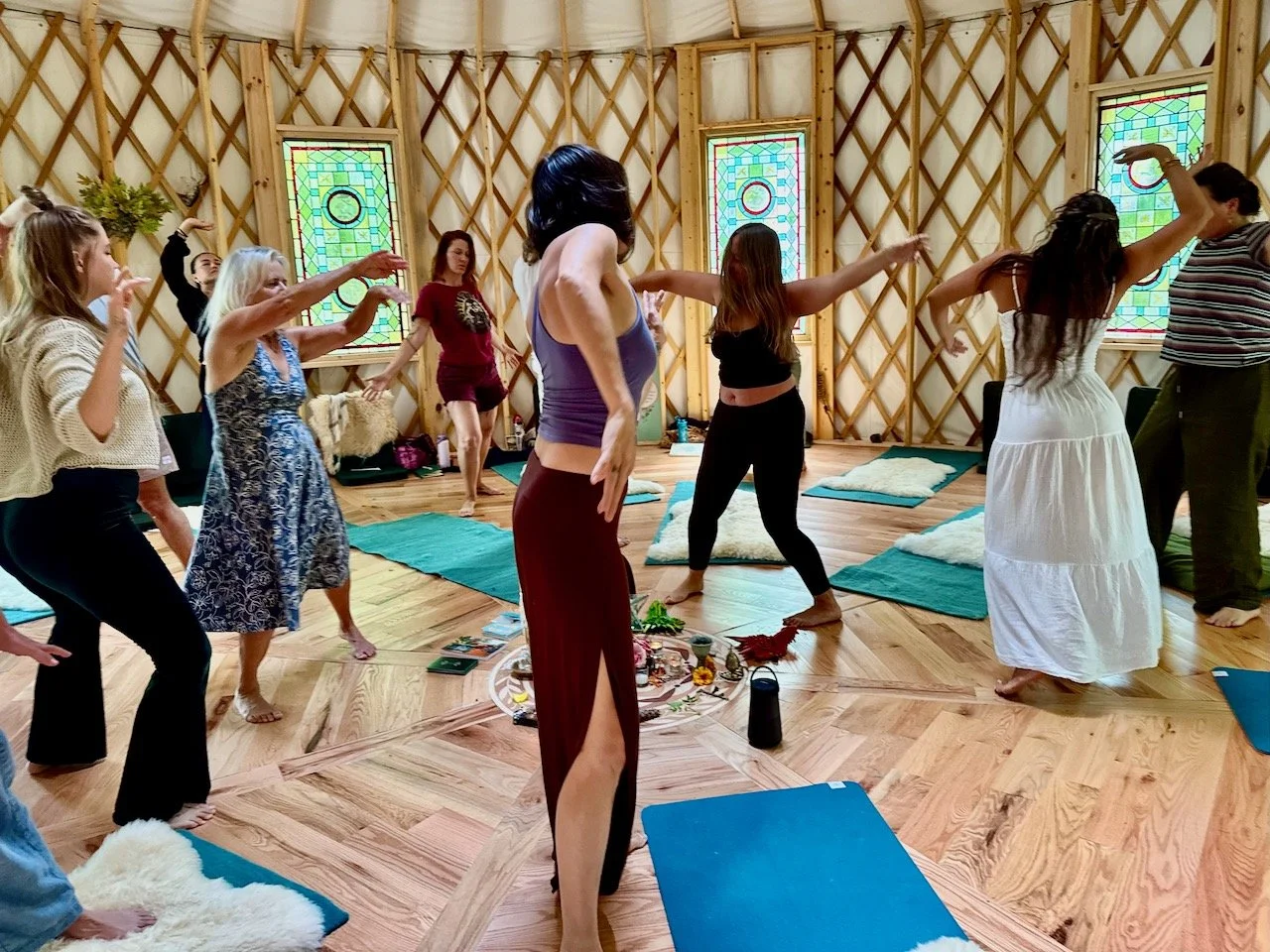 Group of women practicing yoga in a wooden yurt with stained glass windows, some standing on yoga mats, engaged in a dance or movement exercise.
