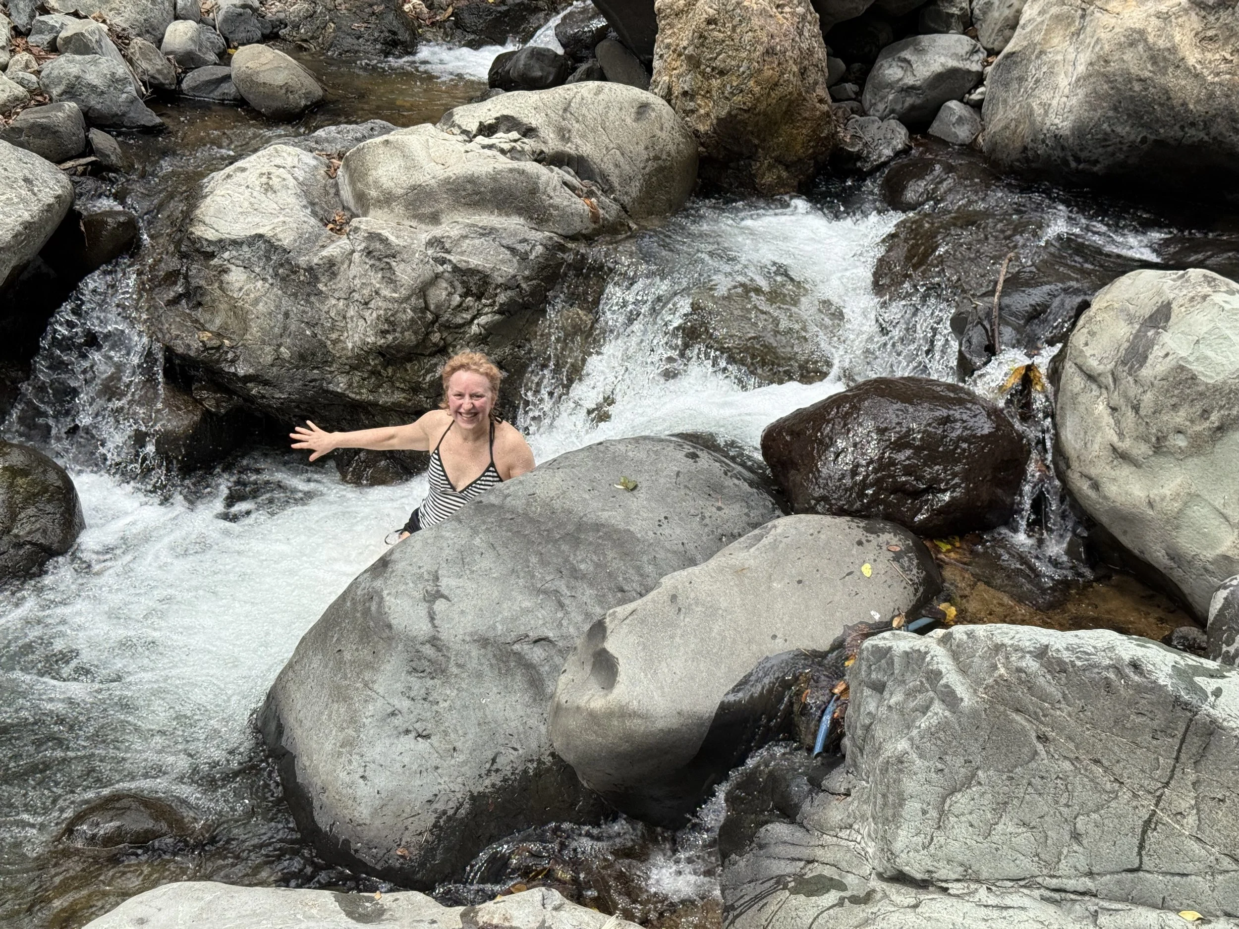 A woman in a striped swimsuit smiling and standing in a rocky river surrounded by large rocks and flowing water.