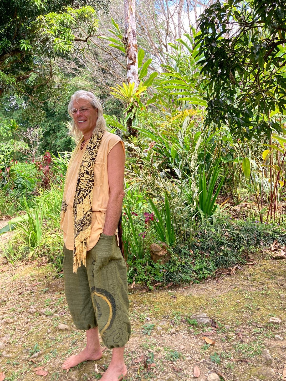A smiling woman with long blonde hair standing barefoot in a lush, green garden surrounded by various tropical plants and trees.