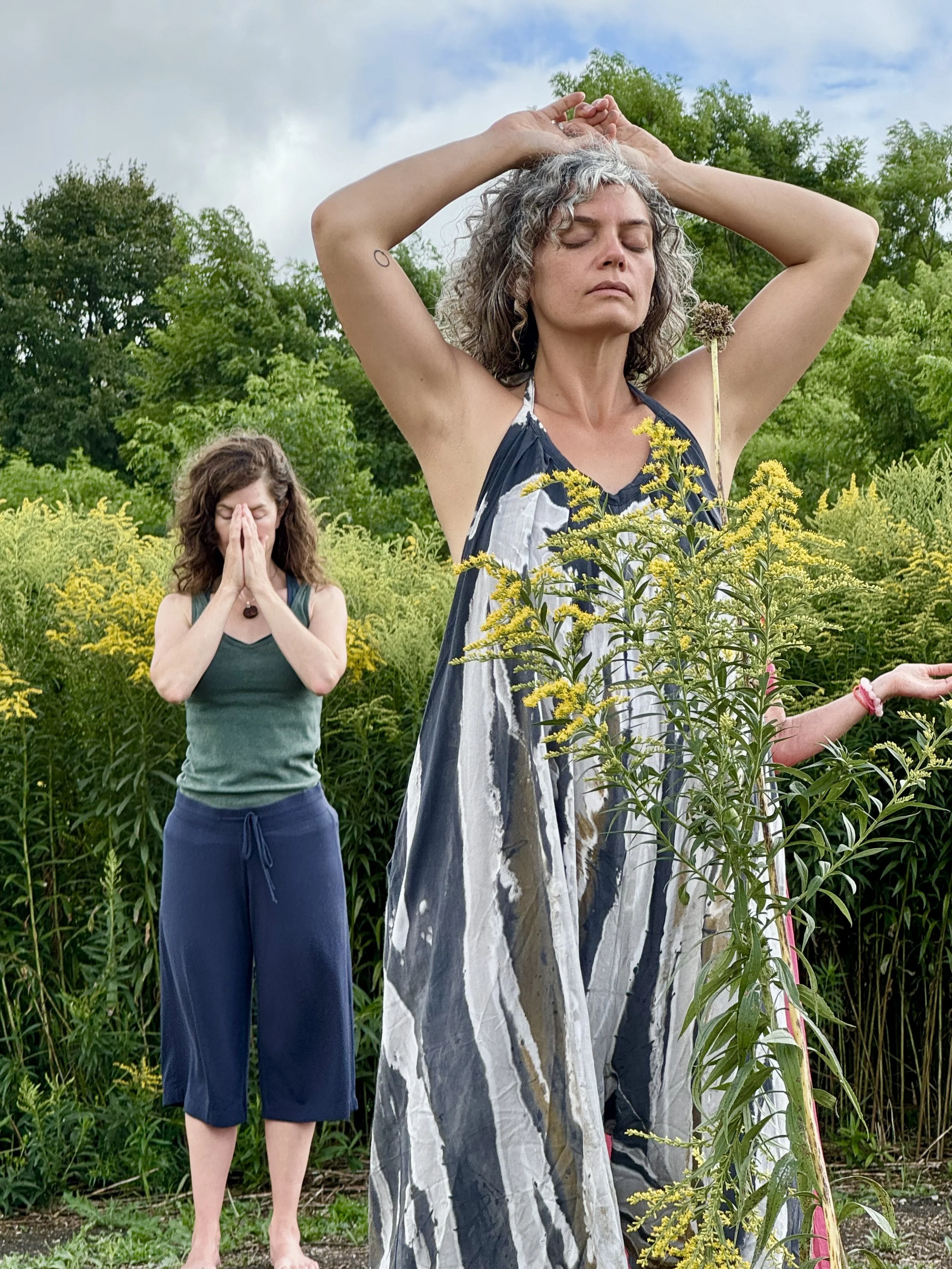 Two women practicing yoga outdoors among greenery and yellow flowers, with eyes closed and hands in prayer or meditation position.