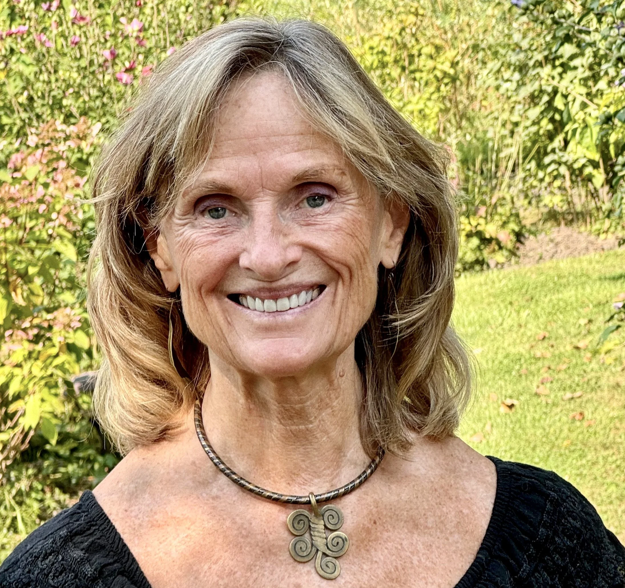 A smiling middle-aged woman with shoulder-length wavy hair, wearing a black top and a necklace with a decorative pendant, standing outdoors with greenery and pink flowers in the background.