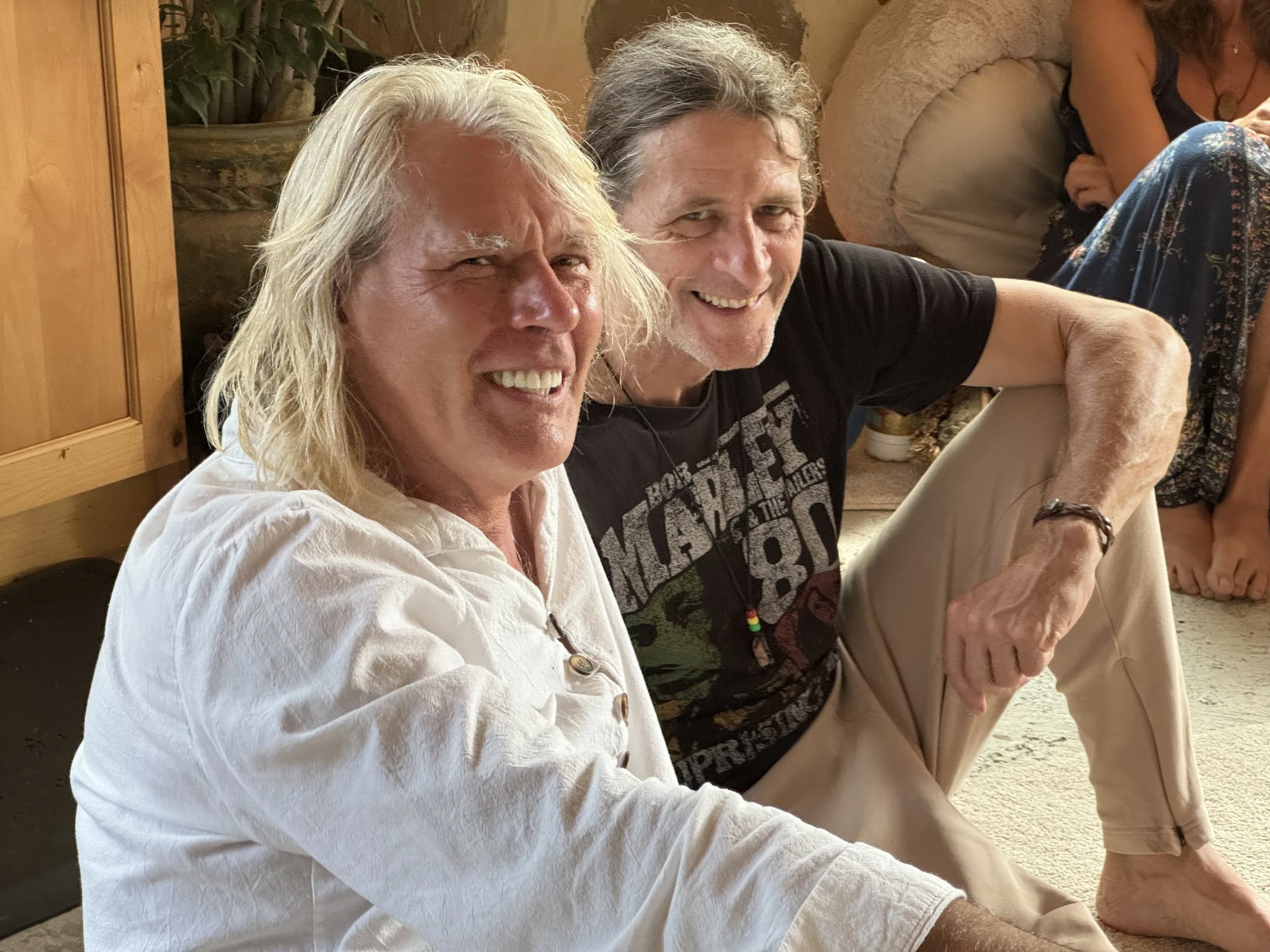 Two smiling men sitting on the floor indoors with a woman and others visible in the background.