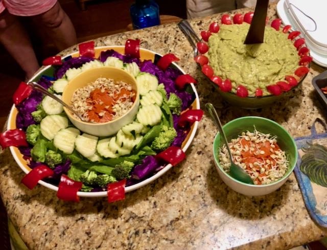 A large vegetable salad with chopped cucumbers, broccoli, and purple cabbage, topped with crushed nuts, served with bowls of shredded cheese and shredded carrots, and a bowl of guacamole garnished with cherry tomatoes.