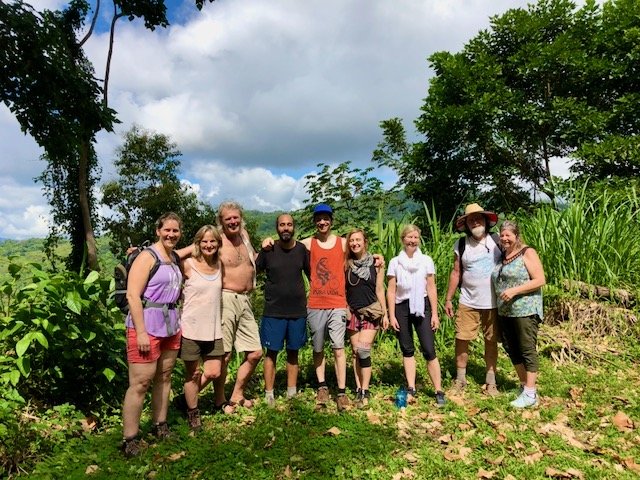 Group of nine people standing outdoors in a lush green, rural setting with trees and plants, on a cloudy day.