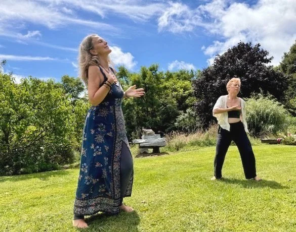 Two women practicing yoga outdoors on a sunny day, surrounded by green trees and grass.