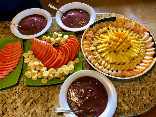 Assorted fresh fruit platter with sliced bananas, papaya, pineapple, and mandarin oranges, along with three bowls of berry sauce or jam on a granite countertop.