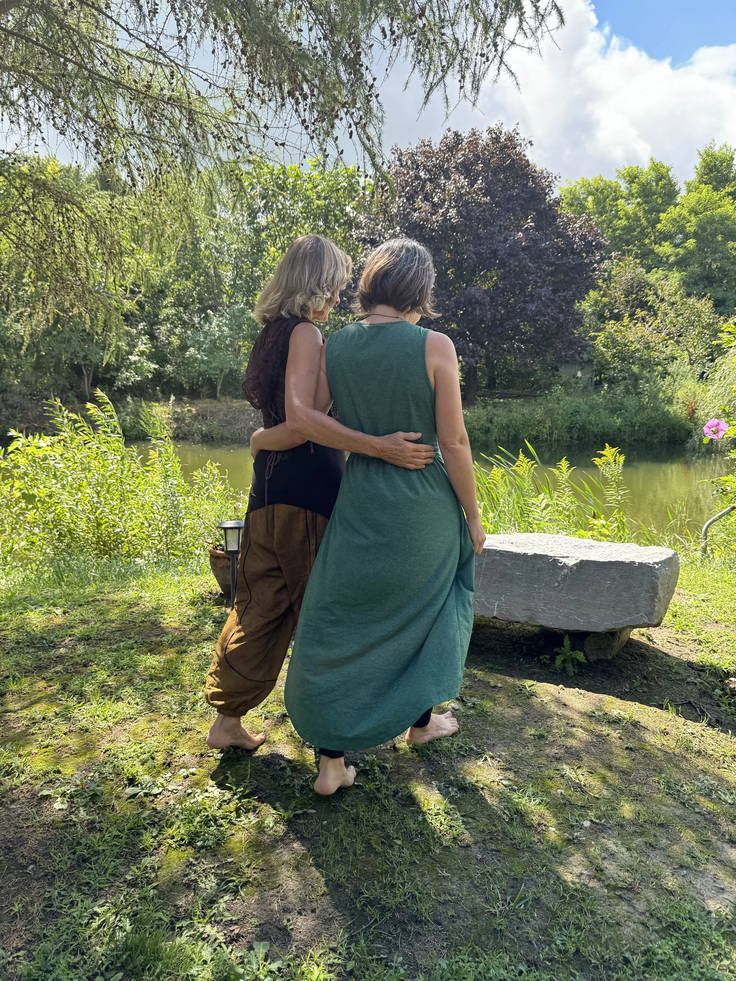 Two women standing close together outdoors by a pond, with one woman's arm around the other's waist, observing the water surrounded by trees and greenery.