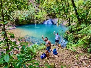 People near a small water body surrounded by lush green trees, with backpacks on the ground, one person waving.