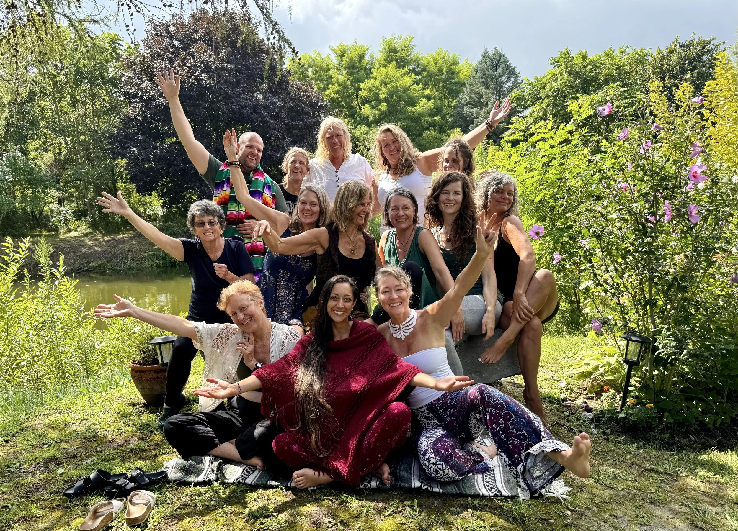 Group of people gathered outdoors near a pond, smiling and posing with arms raised and making peace signs, surrounded by greenery and pink flowers.