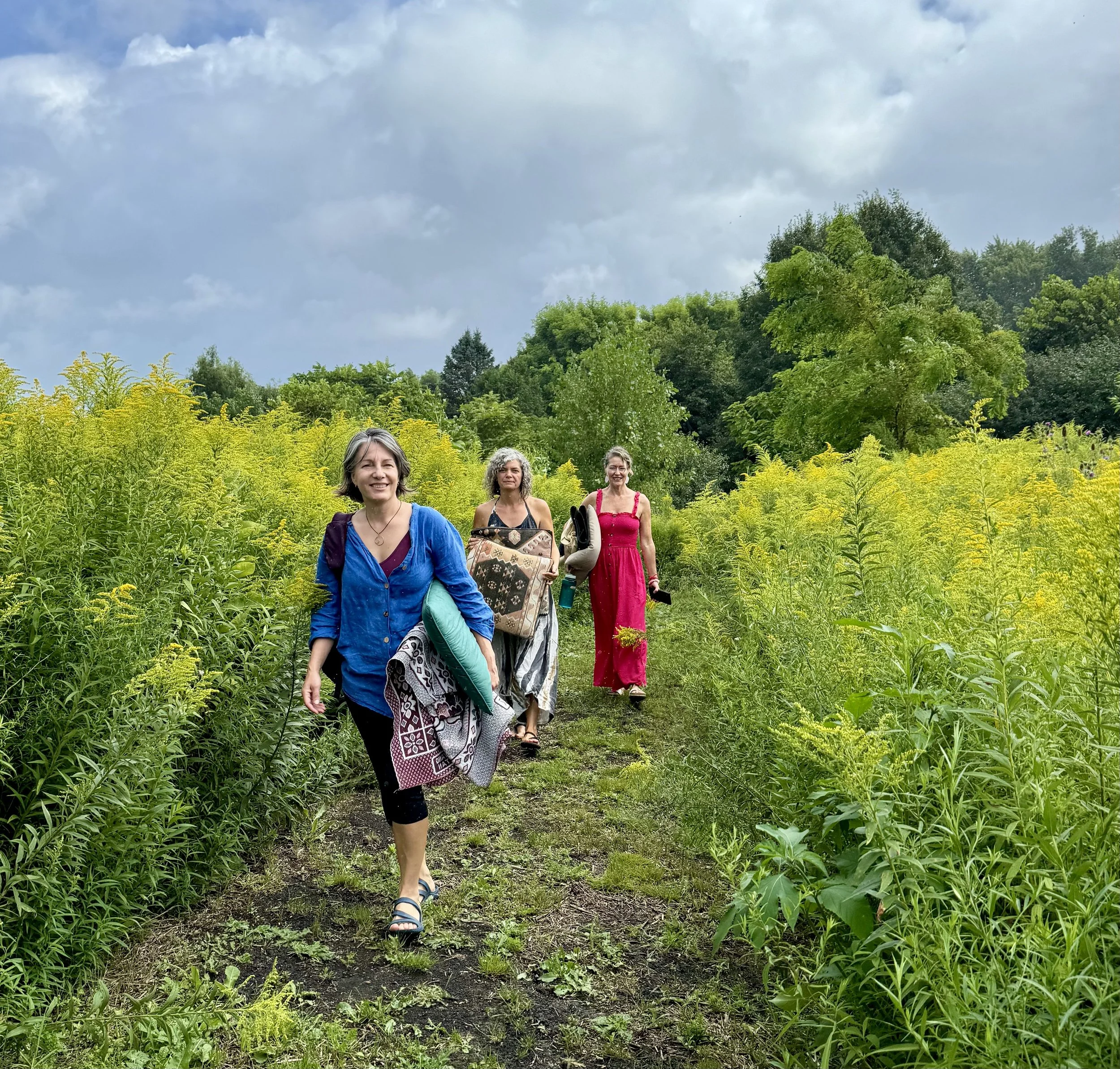 Three women walking along a trail through a lush green field with trees in the background under a cloudy sky.