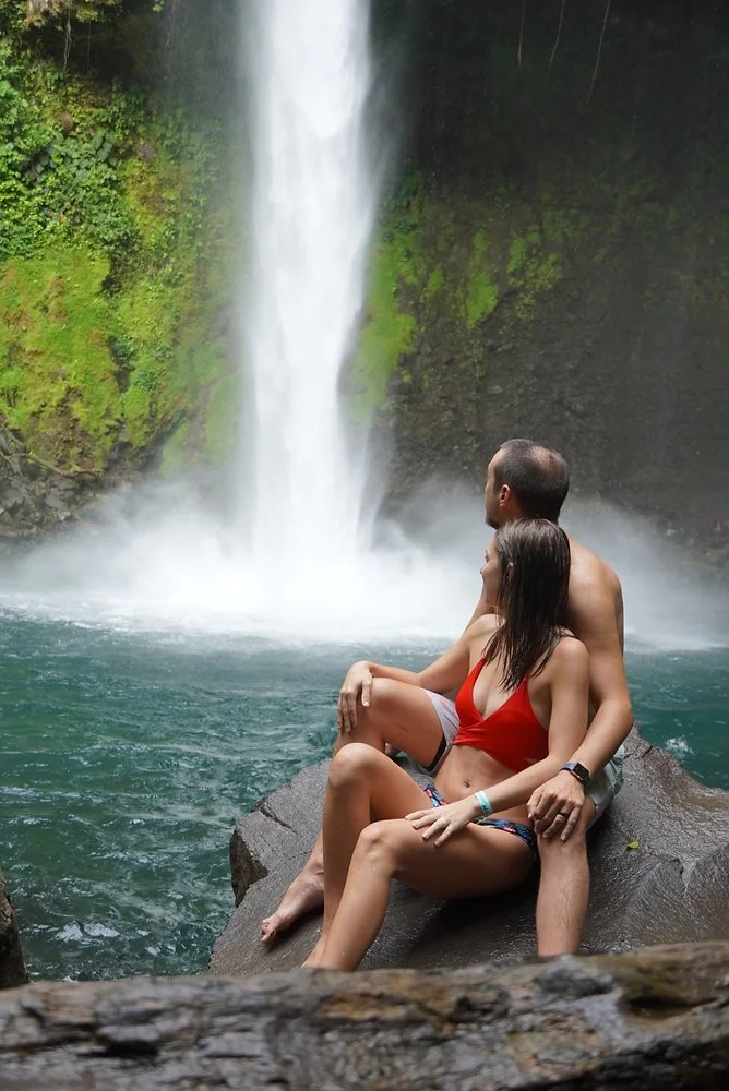 A couple sitting on a large rock in front of a waterfall, with lush green moss-covered rocks and water surrounding them.