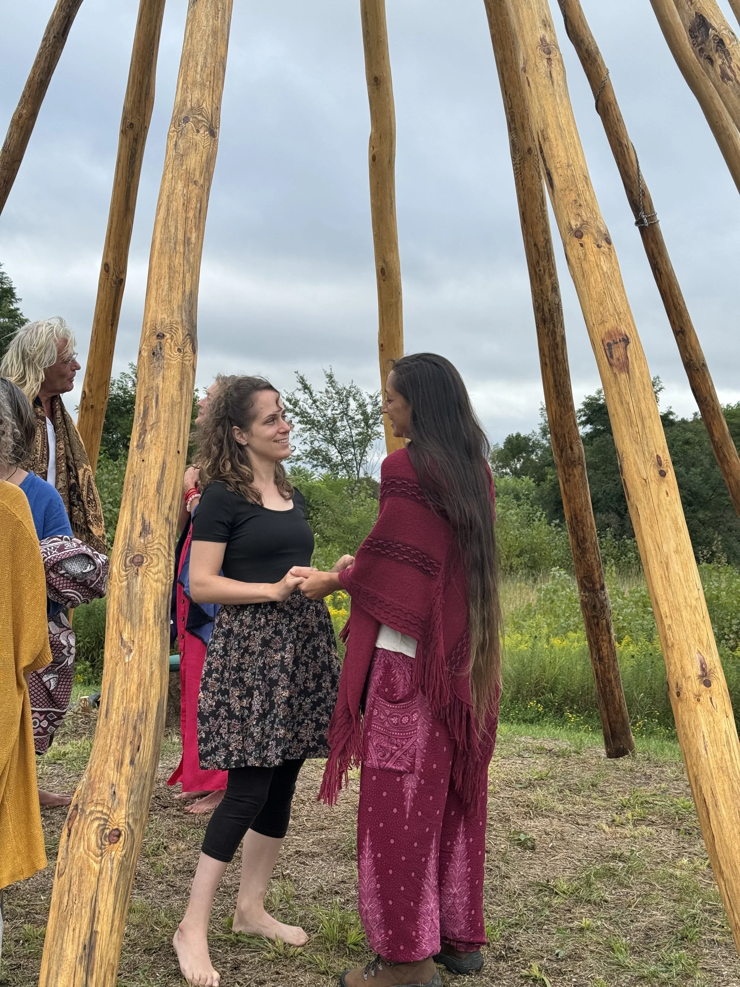 Two women stand hand-in-hand inside a large wooden teepee, smiling at each other, with others nearby outdoors in a grassy field under cloudy skies.