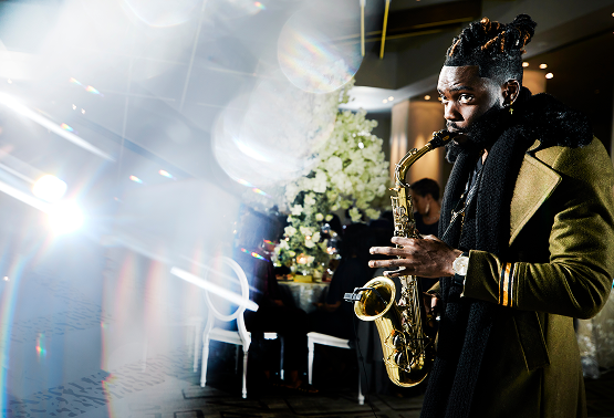 A man playing a gold saxophone at an indoor event, with a floral arrangement and people in the background.