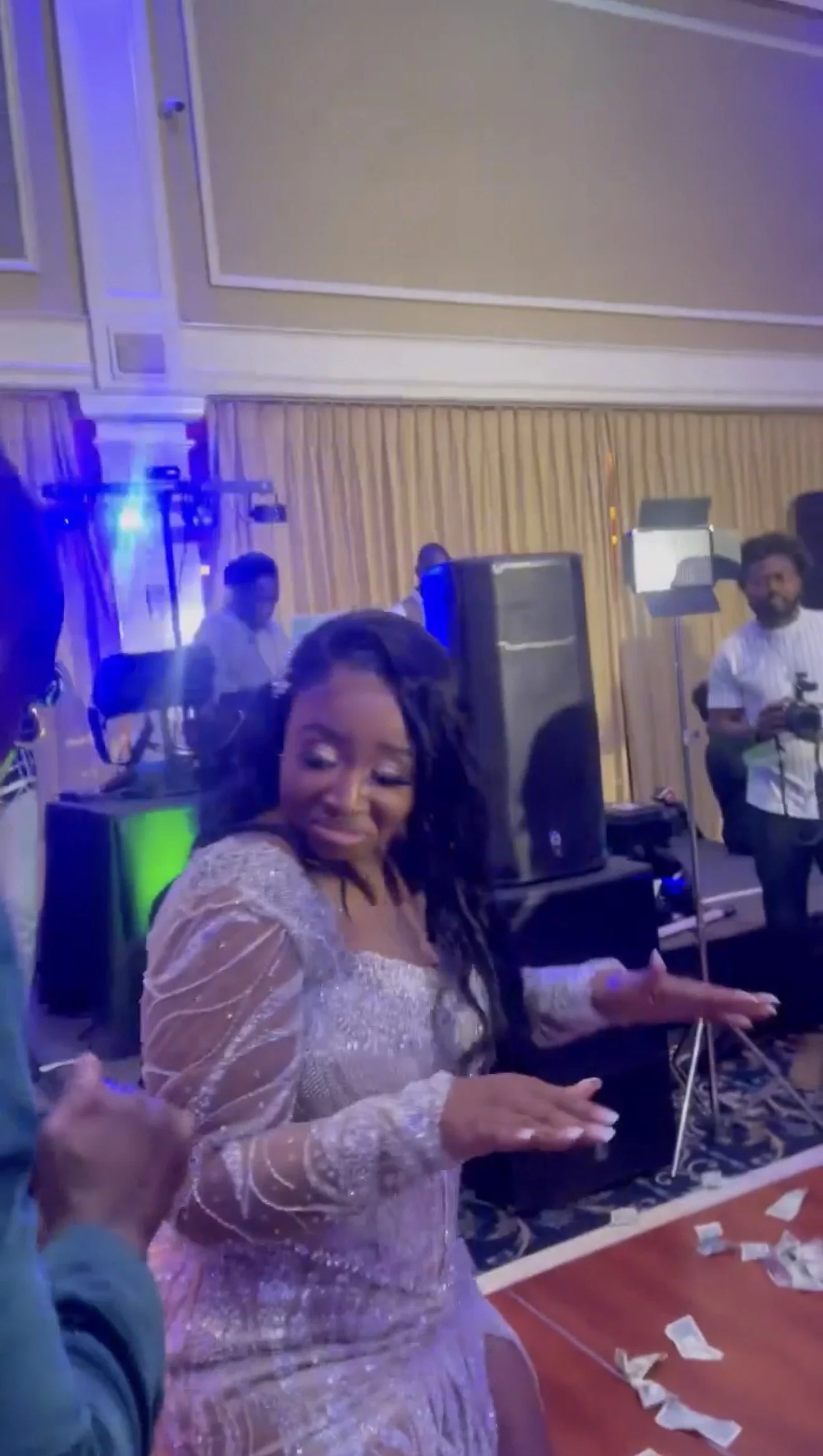 A woman in a sparkly light-colored dress is dancing and smiling at a celebration event. In the background, there are people, DJ equipment, and scattered money on a table.