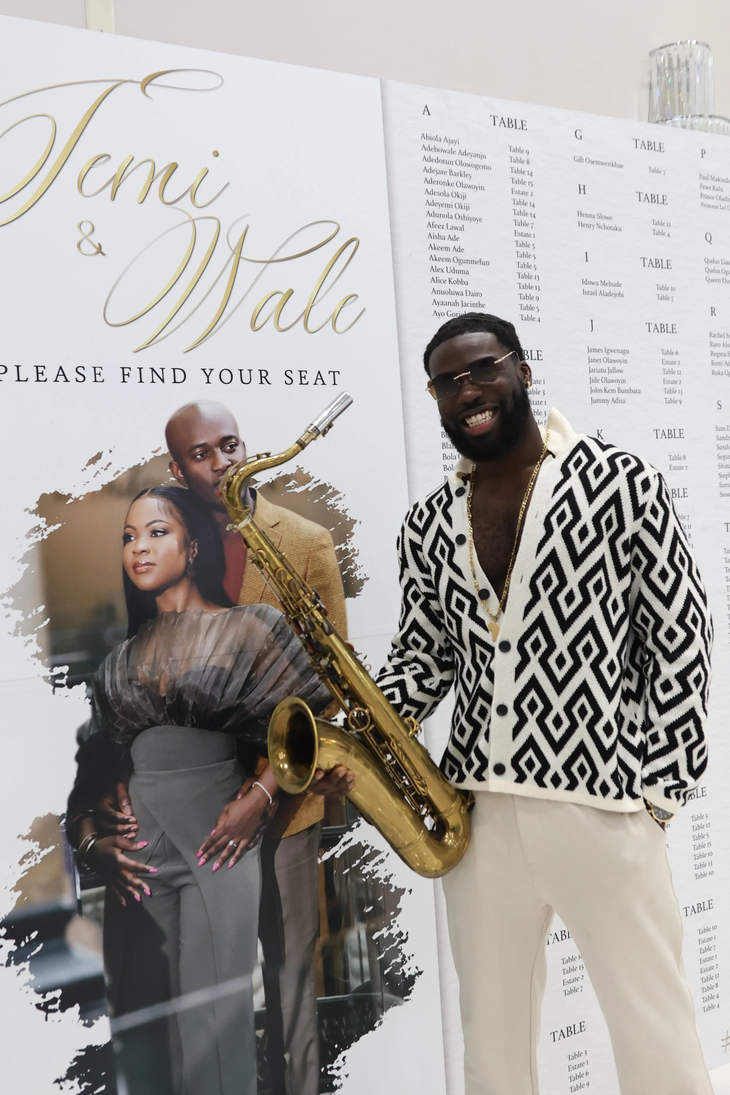 A man standing beside a wedding sign with gold lettering that says "Jemi & Wale" and a table seating chart. The man is smiling, wearing sunglasses, a patterned black and white shirt, and cream pants, and holding a gold saxophone.
