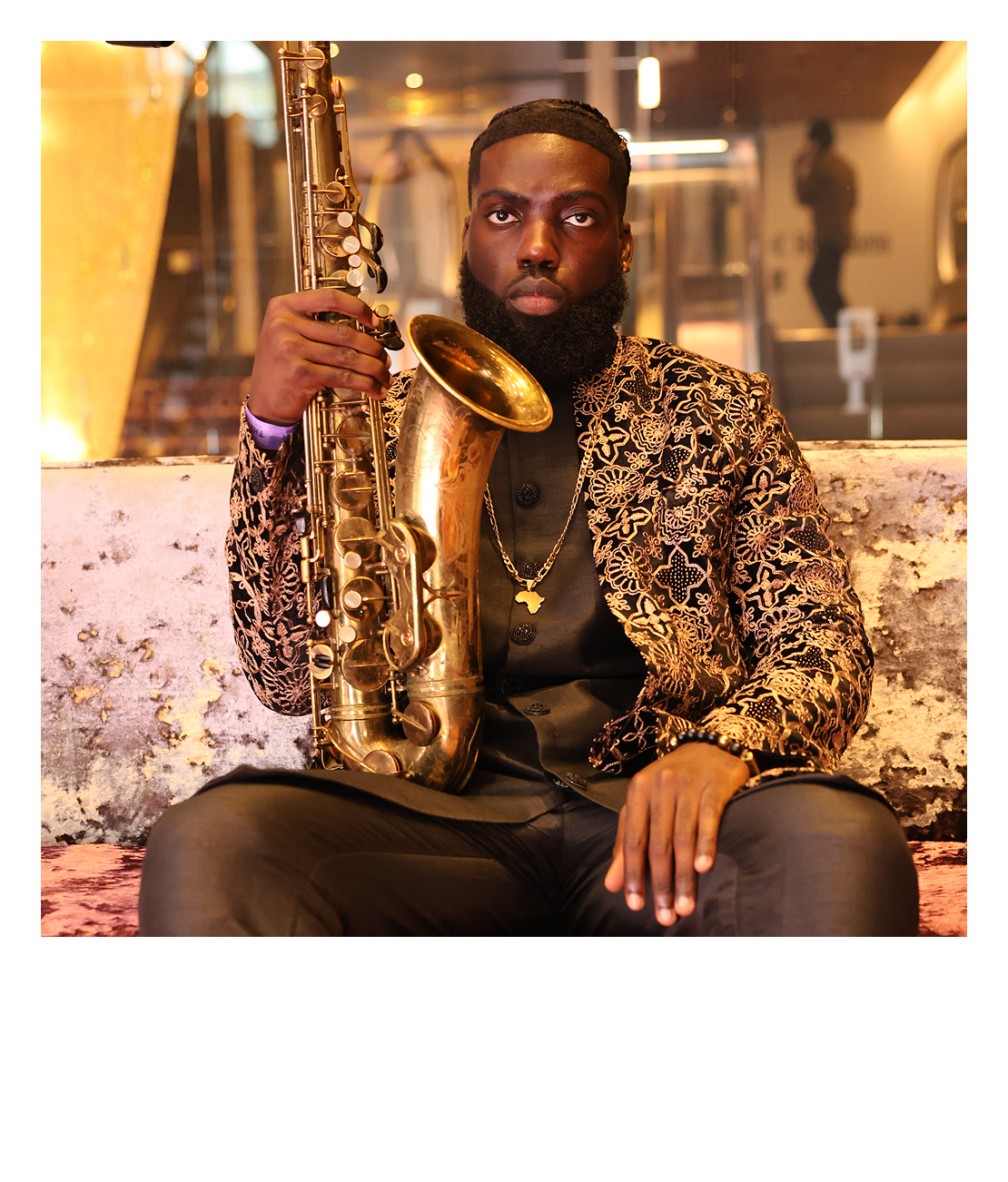 A man wearing a black shirt with a gold embroidered blazer, gold chain, and holding a gold saxophone while sitting on a pink and white couch in a warmly lit modern indoor setting.