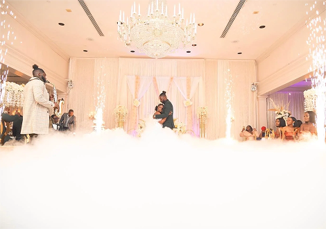 Couple dancing on a decorated wedding stage with chandeliers and cloudy fog, surrounded by guests.
