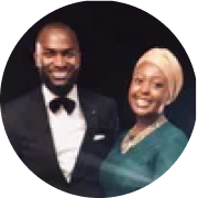 A smiling man in a black tuxedo with a bow tie and a woman in a teal dress and beige headwrap, both posing for a photo against a dark background.