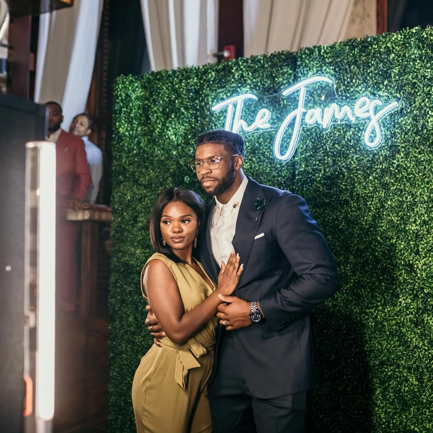 A man and woman posing together at a social event in front of a green hedge backdrop with a neon sign that reads 'The James'.