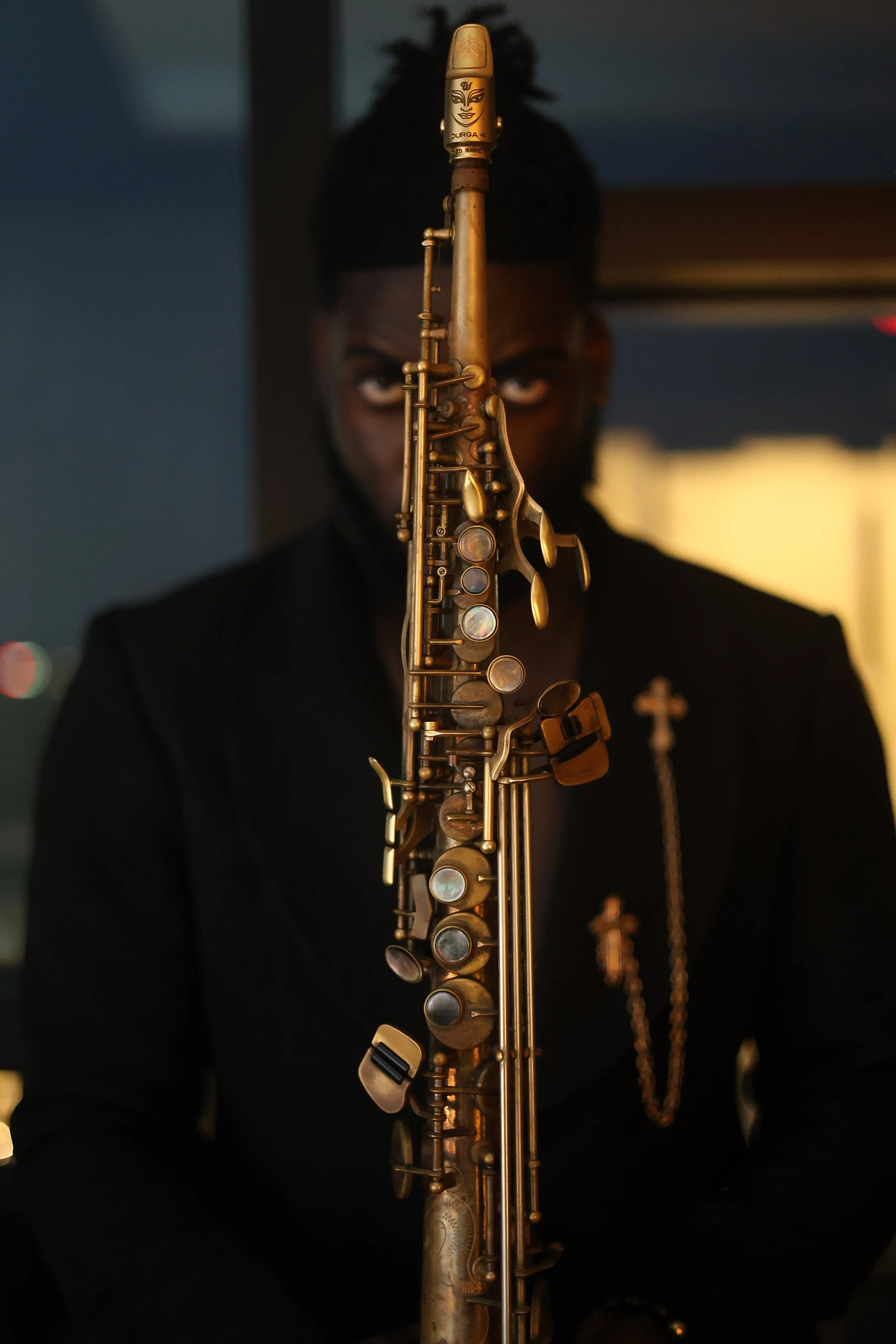 A person in a dark suit holding a gold saxophone with a face-shaped mouthpiece, facing forward with eyes visible behind the instrument.