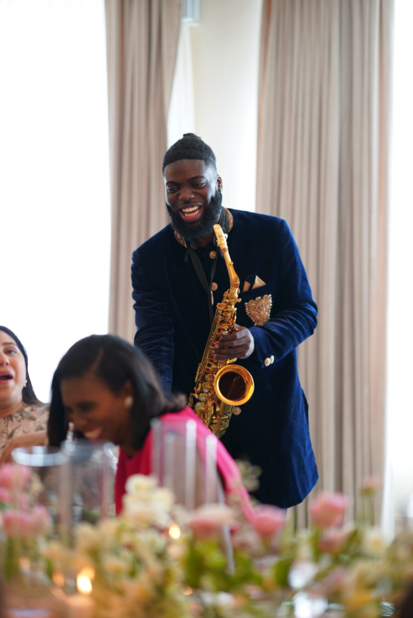 A man with a beard and dark hair in a bun, wearing a navy blue velvet jacket with gold embroidery, playing a saxophone at a social gathering with a blurred background of a dining area and other people.