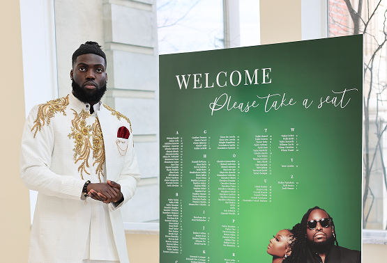 A man in traditional white and gold attire standing next to a green event seating chart that says 'Welcome, Please Take a seat,' in a well-lit room with large windows.