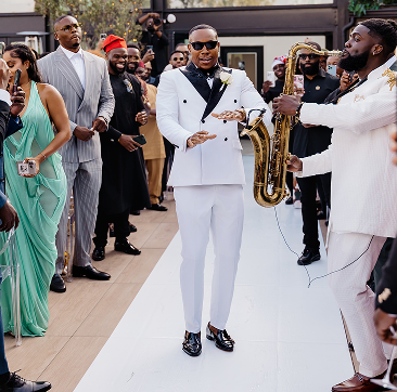A man in a white suit with black shoes and sunglasses holding a microphone, standing on a white carpet at a celebration event with multiple guests, some wearing formal attire, and a musician playing a saxophone.