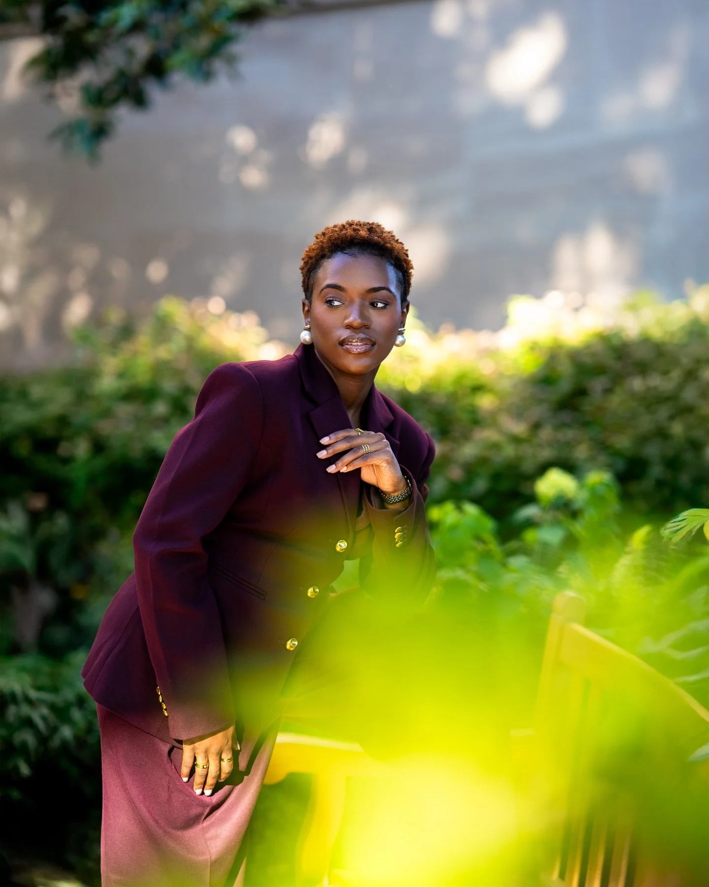A woman with short curly hair wearing a dark maroon blazer and matching skirt, standing outdoors among green foliage, with a blurred background of trees and light.