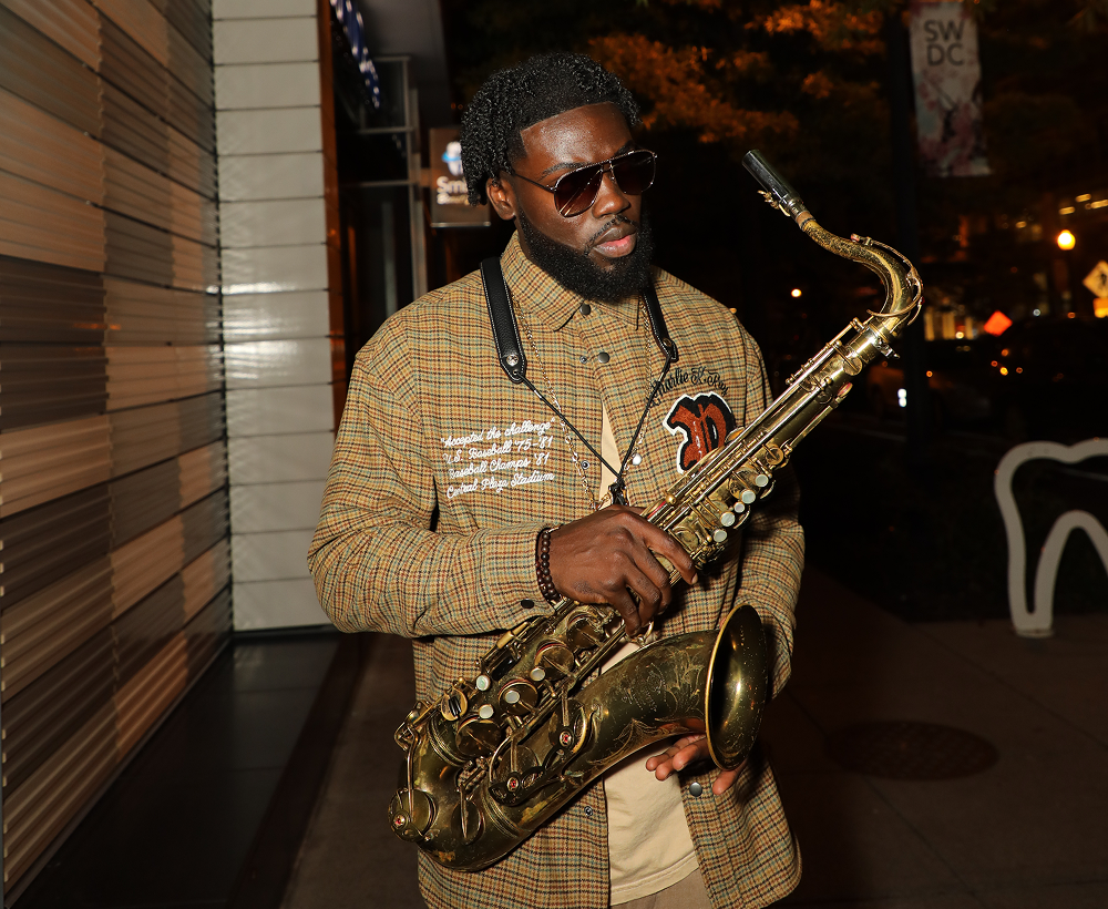A man with sunglasses and a plaid jacket holding a saxophone on a city street at night.