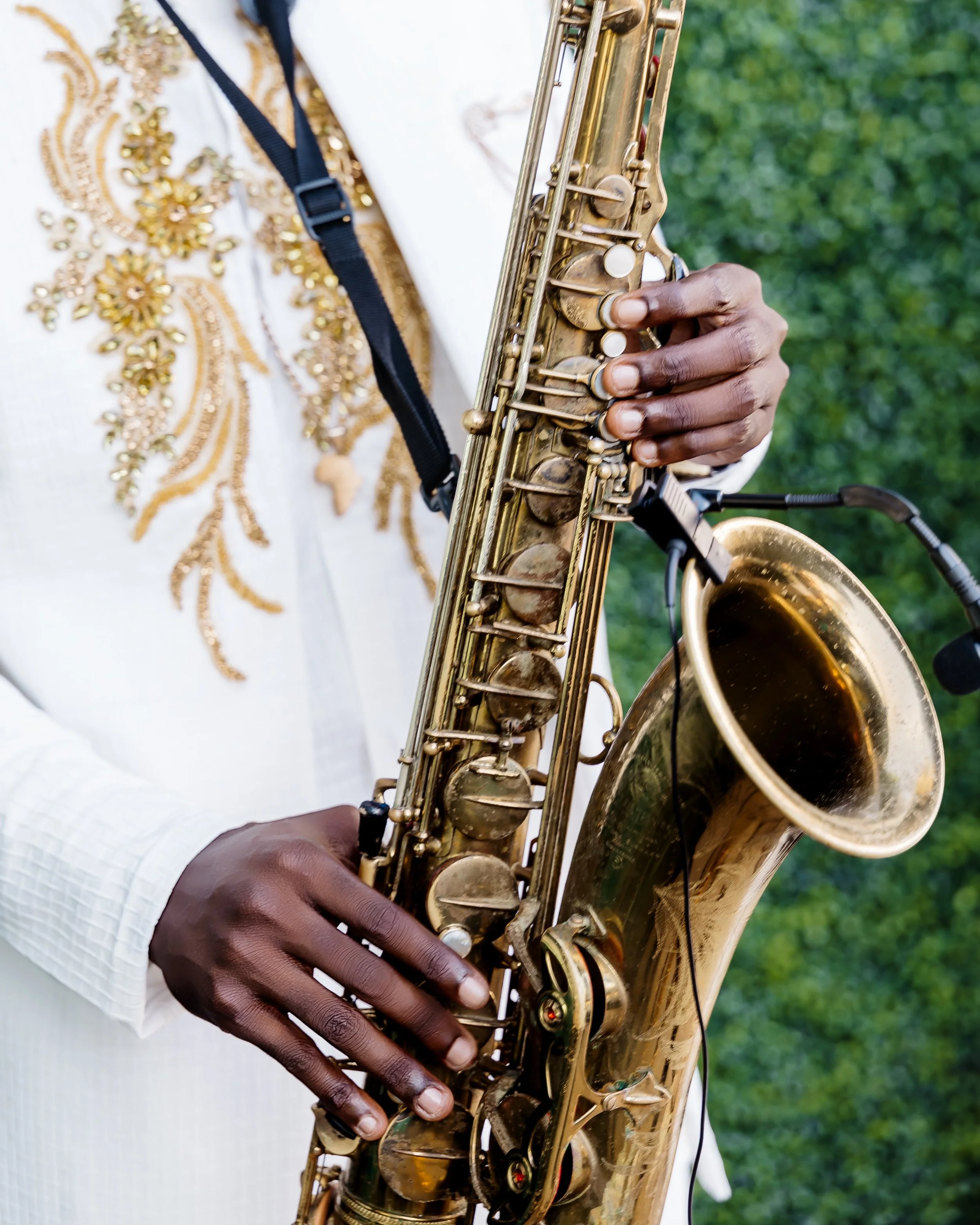 Close-up of a person playing a gold saxophone outdoors, wearing a white garment with gold embroidery, holding the instrument with one hand, green foliage in the background.
