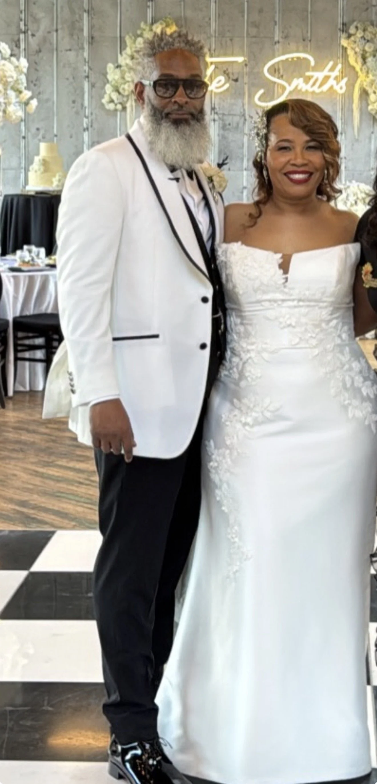 A man and woman dressed in wedding attire standing indoors. The man wears a white tuxedo jacket with black trim, black pants, and shiny black shoes. The woman wears a strapless white wedding gown with lace detailing, smiling with red lipstick. The background features floral arrangements and a glowing neon sign.