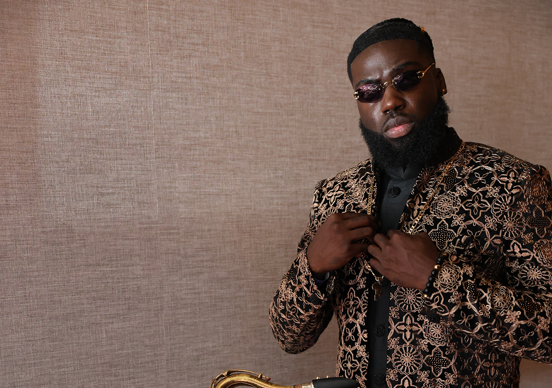 A man with dark hair and a beard wearing sunglasses, a black shirt, and a brocade jacket with a floral pattern, standing against a beige textured wall.
