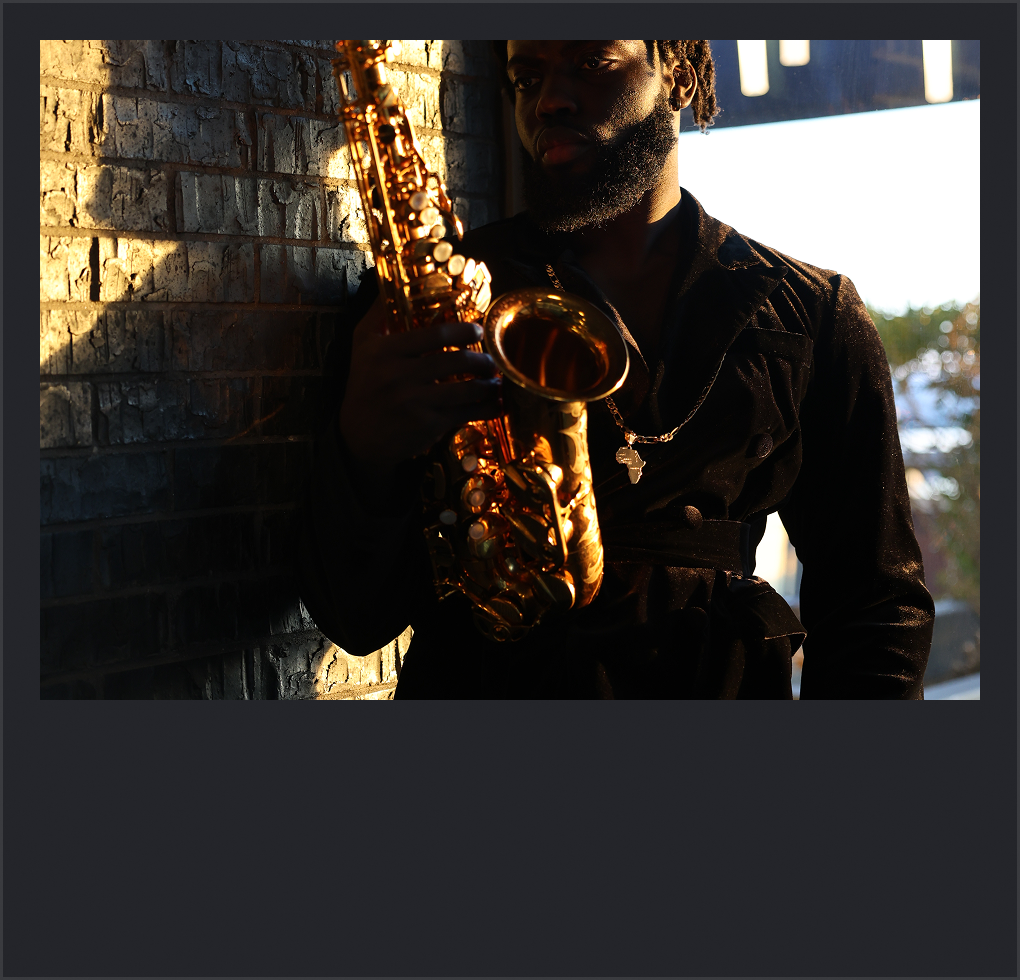 A man with a beard holding a saxophone, standing against a brick wall with sunlight casting shadows on the wall.