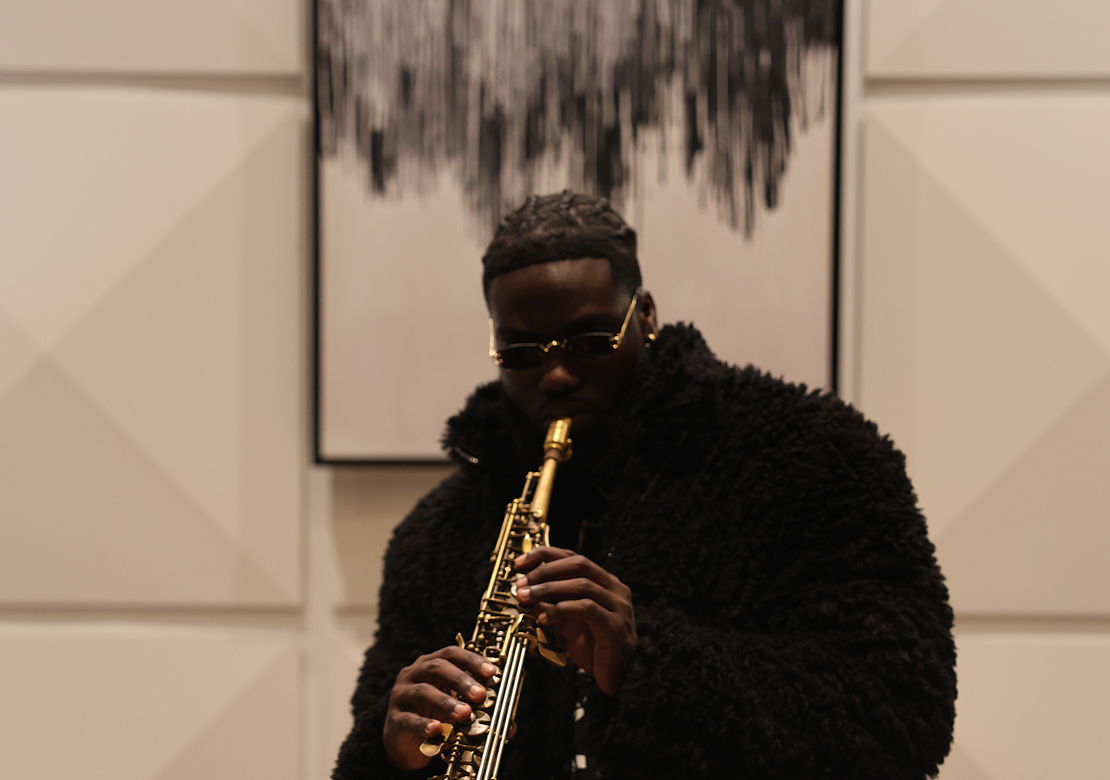 A person wearing sunglasses and a black fuzzy jacket playing a saxophone.