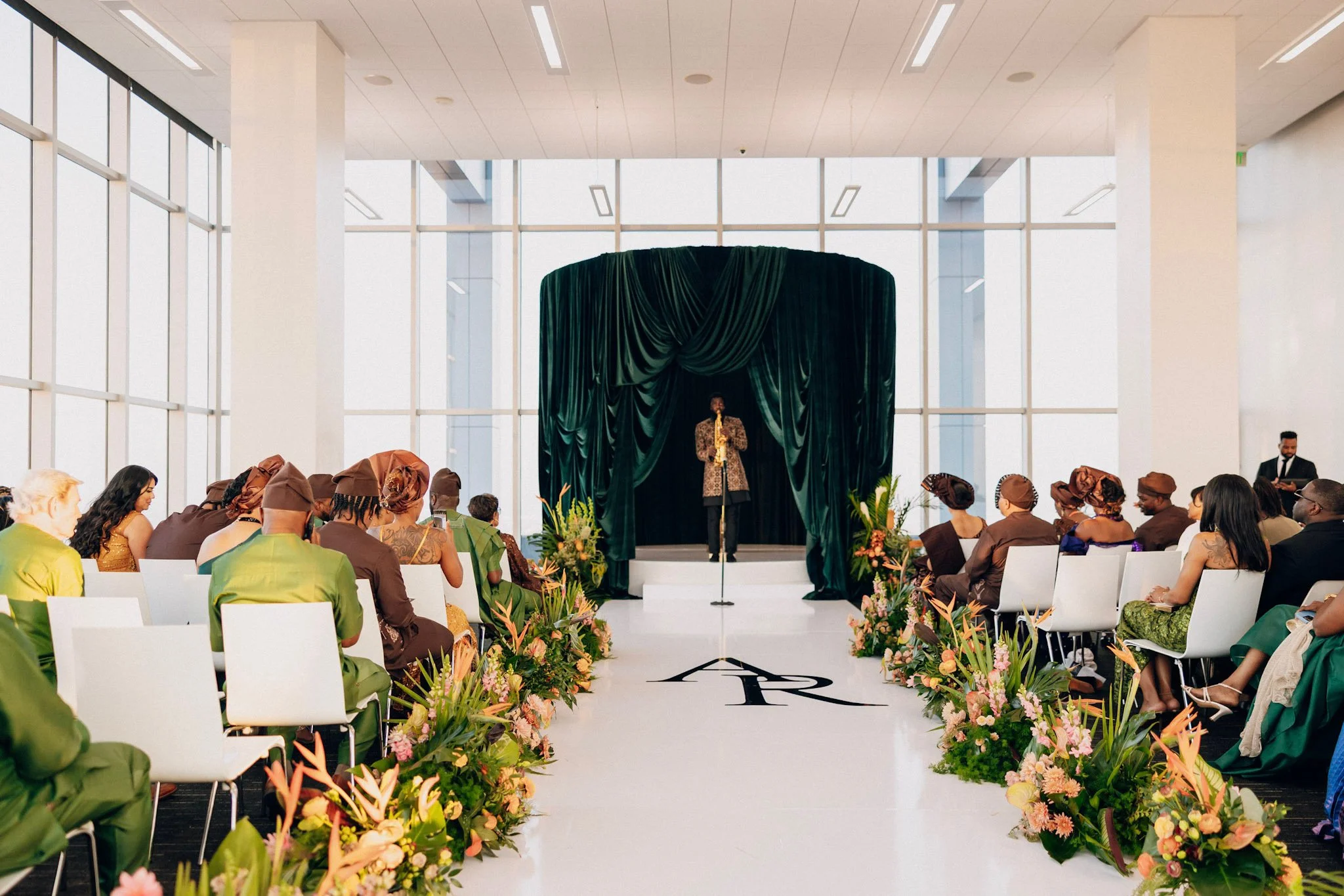 A fashion show with models sitting on both sides of a runway decorated with pink and orange flowers, a performer singing on a stage with green curtains, and a modern, bright interior with large windows.