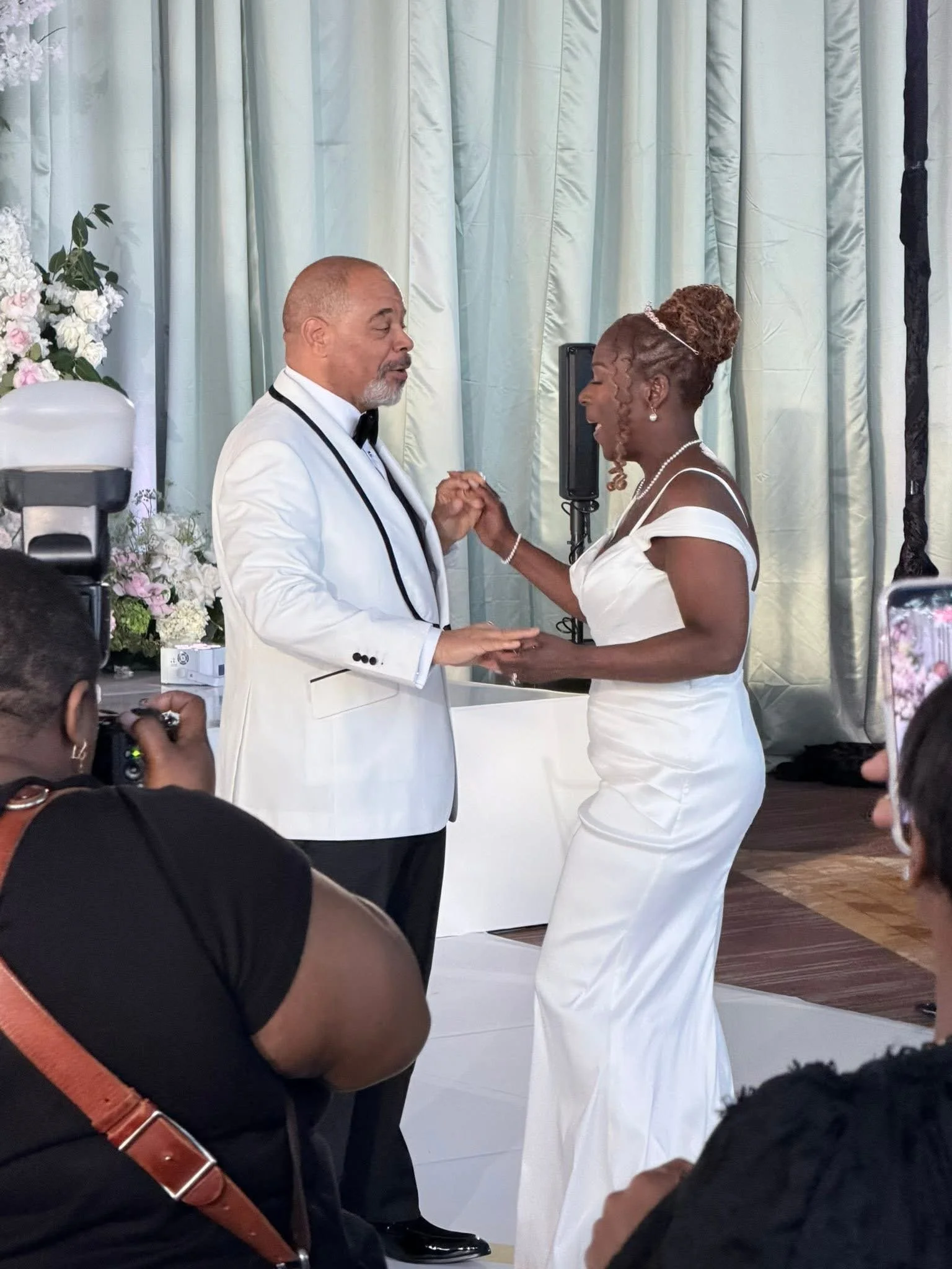 A bride and groom holding hands and dancing during a wedding ceremony. The bride is wearing a white gown and pearl jewelry, and the groom is dressed in a white tuxedo with a black bow tie. They are in front of a light-colored curtain with floral decorations nearby.
