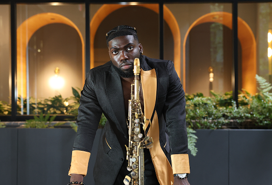 A man standing behind a golden saxophone, looking at the camera, with a backdrop of large archways and indoor plants.