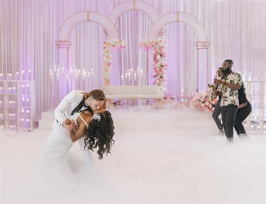 A bride and groom share a dance at their wedding reception, with a photographer capturing the moment in a decorated event space with pink and white floral arrangements, arch decorations, and a fog effect on the floor.