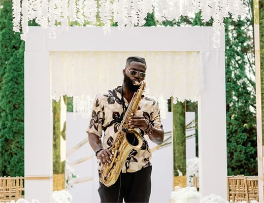 Man playing saxophone at an outdoor event with a white floral arch and greenery in the background.