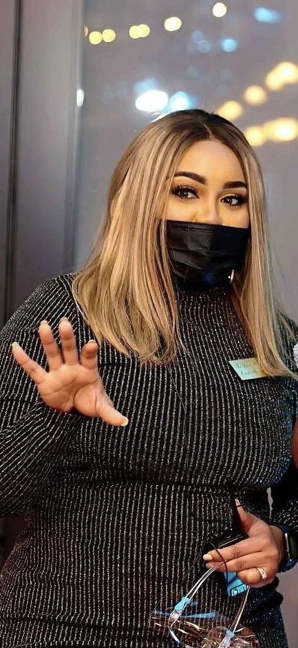 A woman with blonde hair, wearing a black face mask and a black and white striped top, is holding a glass of wine and making a gesture with her hand. She appears to be indoors with city lights visible in the background.