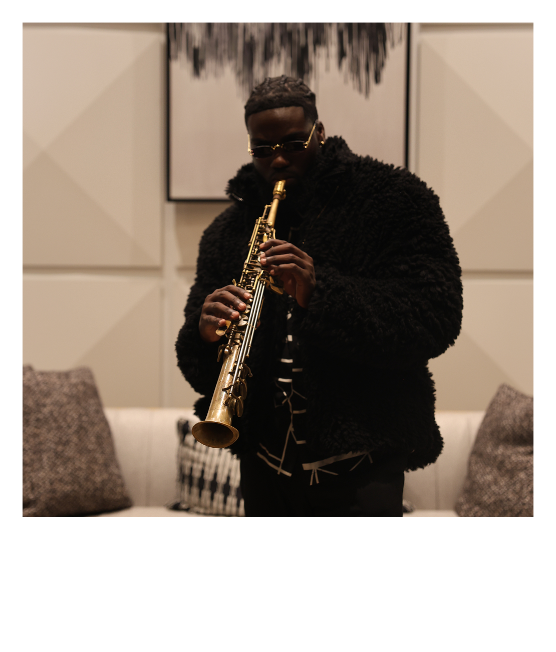 A man wearing sunglasses and a black furry jacket playing a gold-colored soprano saxophone indoors, with abstract artwork on the wall behind him.