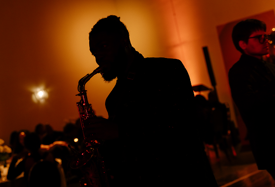 Silhouette of a person playing a saxophone at a dimly lit event with a warm, orange background.