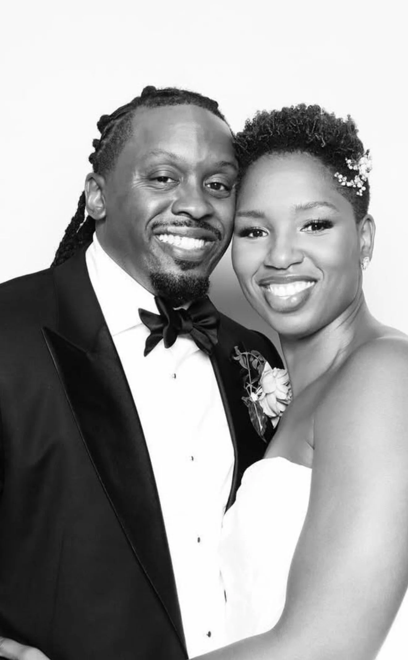 Black and white photo of a smiling man in a tuxedo and a woman in a strapless dress, close together and facing the camera.