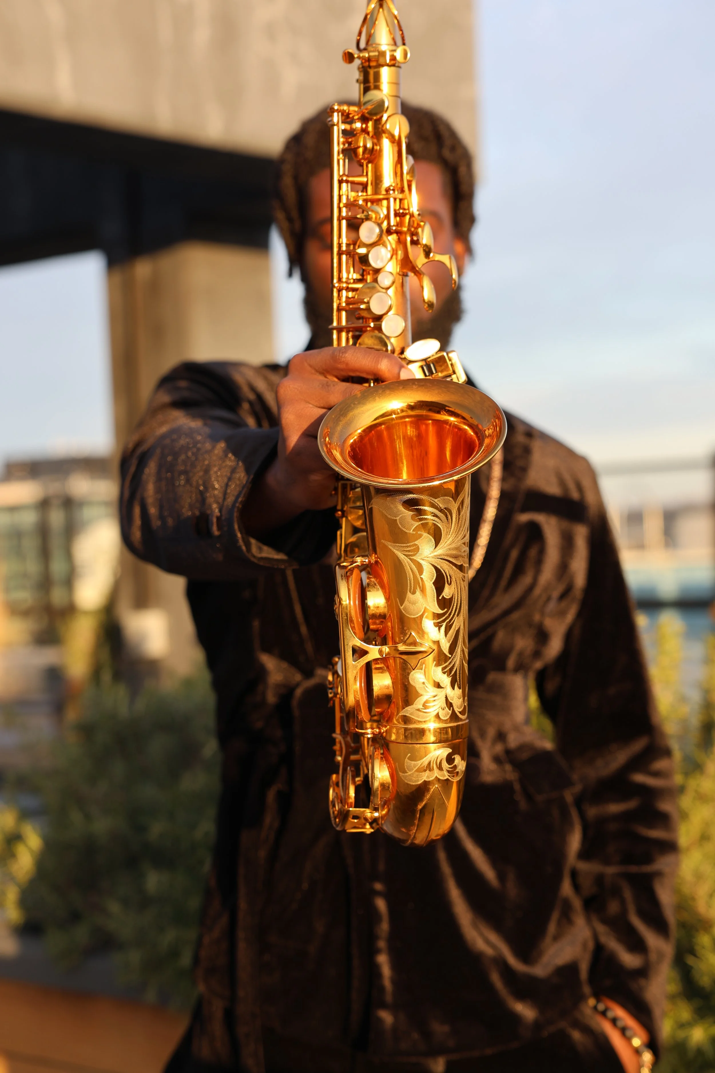 Person playing a gold saxophone outdoors with blurred cityscape background during golden hour.