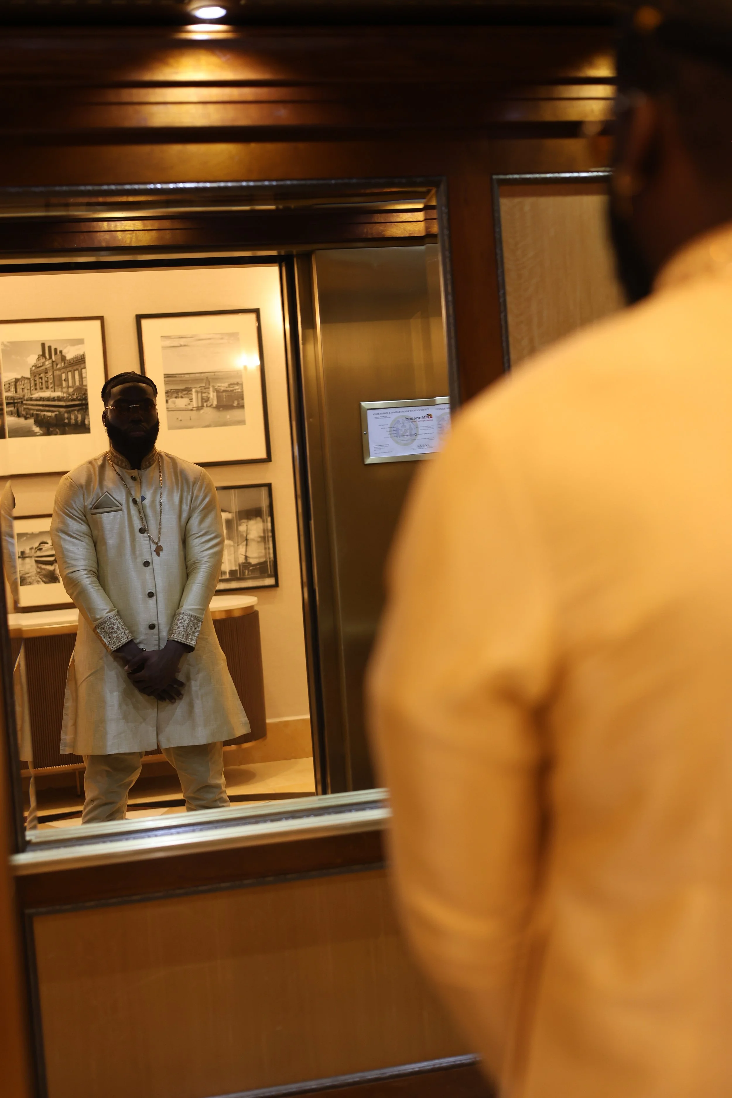 A man wearing traditional Indian attire and jewelry is looking into a mirror, with framed black and white photographs on the wall behind him. A person in a yellow shirt is partially visible in the foreground, looking at him.