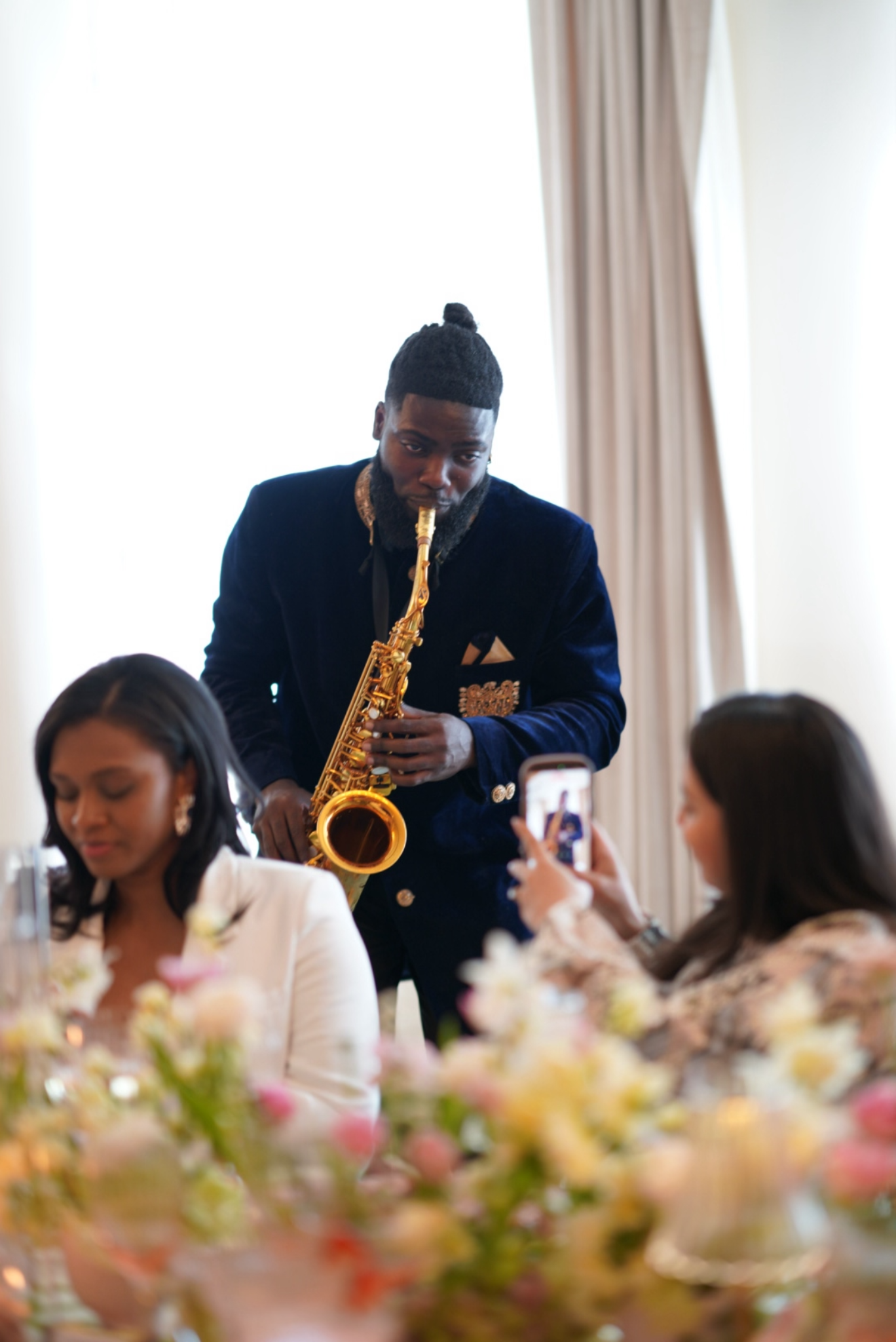 Man with a guitar at a formal event, sitting at a table with women, with flowers in the foreground.